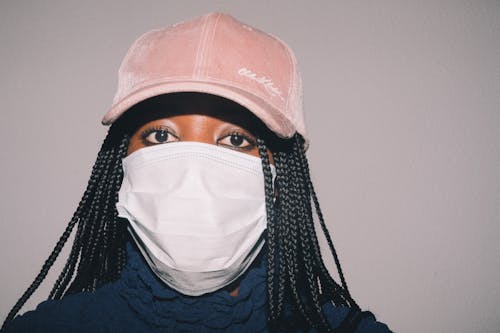 Adult African American woman in casual clothes and pink cap and medical mask while standing against gray background