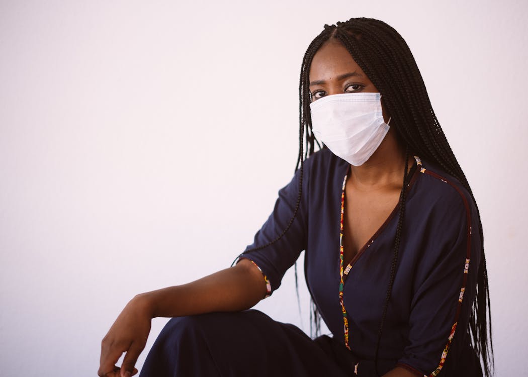 Free Side view of unrecognizable African American woman in casual dress and protective mask sitting against gray background looking at camera Stock Photo
