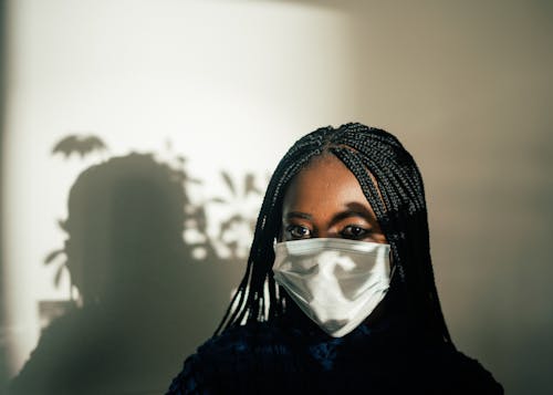 Unrecognizable African American female in medical mask against white wall with shadow of leaves plant on face