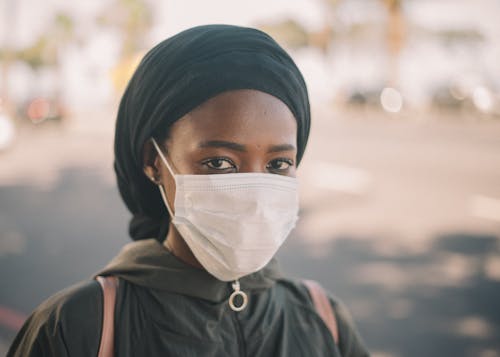 Unrecognizable young ethnic woman in jacket and headscarf and protective mask standing on city street