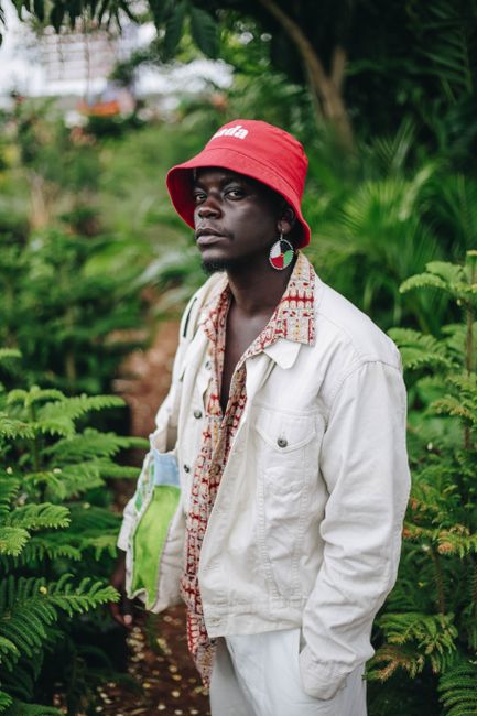 Elegant man in a bucket hat and jacket poses among green foliage in an outdoor setting, showcasing urban fashion.