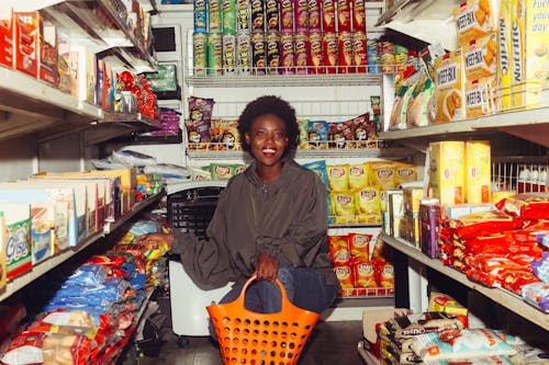 Full length of cheerful ethnic woman in casual clothes with shopping bag squatting among store shelves choosing food for buying