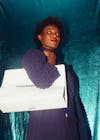Free Low angle of serious African American lady in violet dress standing with white shopping bag on shoulder and looking away against metallic blue textile background Stock Photo