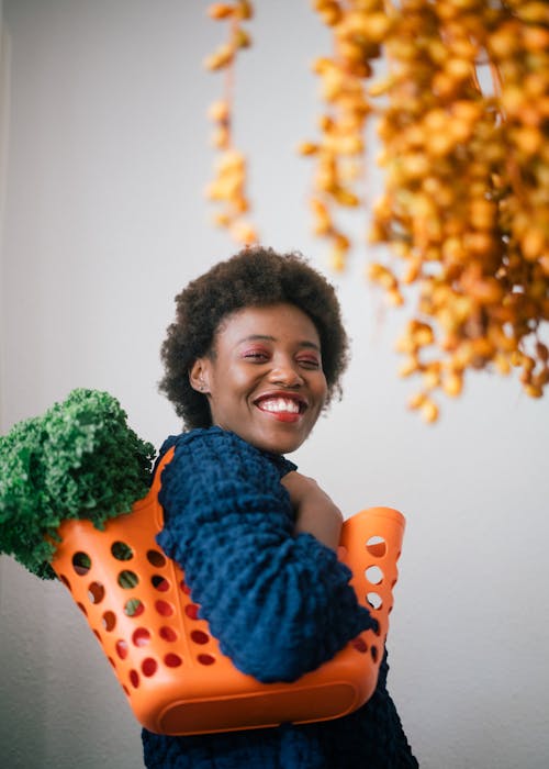 Free Happy young black woman holding basket with lettuce on shoulder and cluster of yellow dates in grocery store on gray background Stock Photo