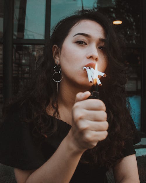Confident young ethnic female with dark curly hair lighting many cigarettes in mouth and looking at camera
