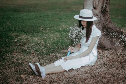 Young woman in a hat and white dress holding flowers, sitting by a tree outdoors.