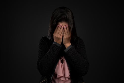 Unrecognizable sad female with dark hair in casual clothes covering face with hand against black background