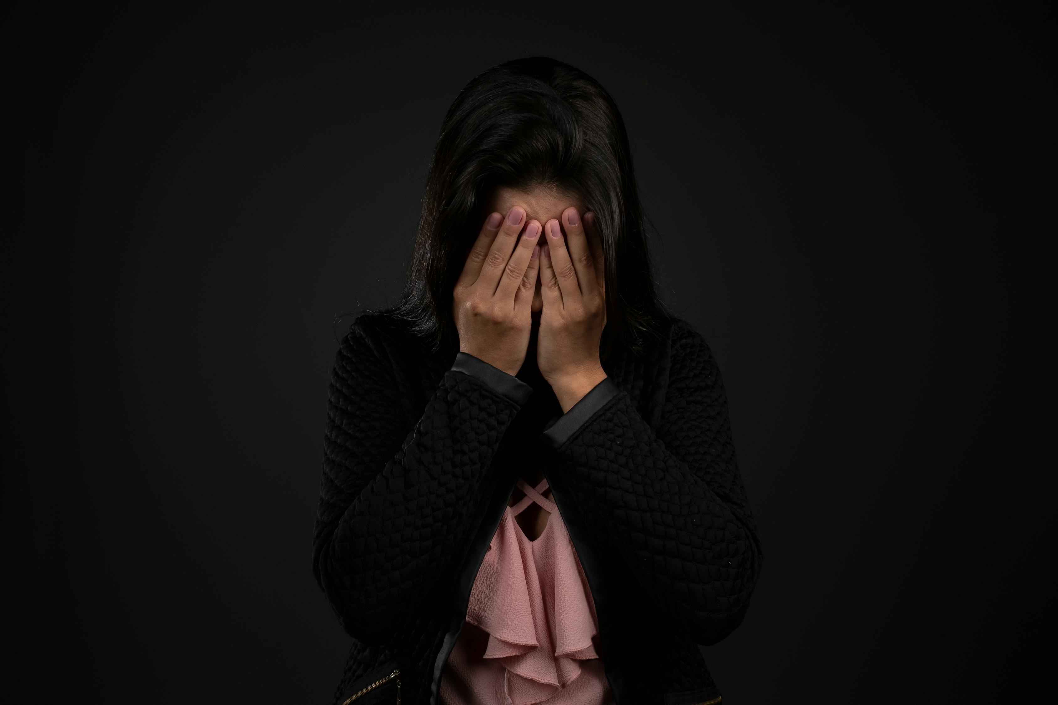 Unrecognizable sad female with dark hair in casual clothes covering face with hand against black background