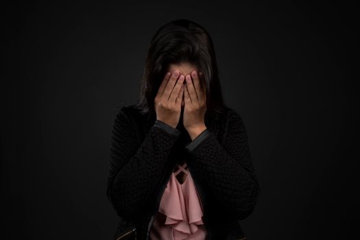 一位深色头发、穿着休闲服的悲伤女性，在黑色背景前用手遮住了脸