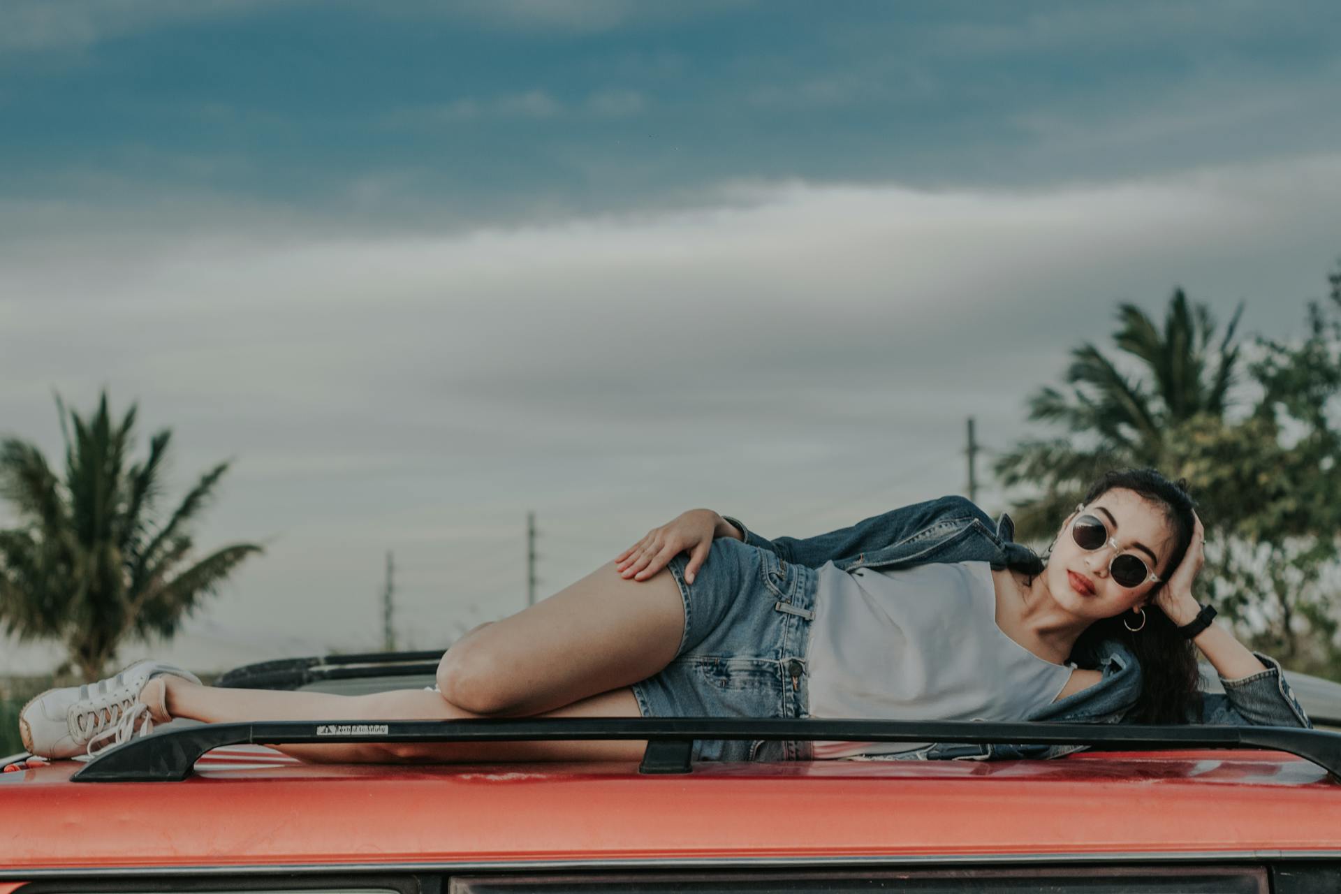 A fashionable woman in denim poses confidently on a car roof under a clear sky.