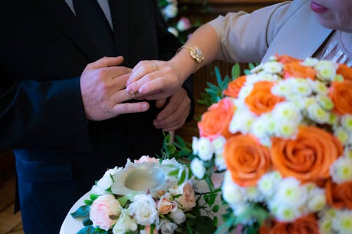 Unrecognizable groom with bouquet putting ring on finger of anonymous groom while standing near table with flowers during wedding ceremony