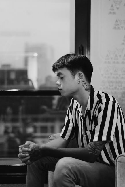 Black and white side view of teen guy with tattoos wearing striped shirt sitting near big window in comfortable armchair in room