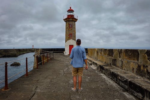 Back view of anonymous traveler standing on stone pier and looking at lighthouse