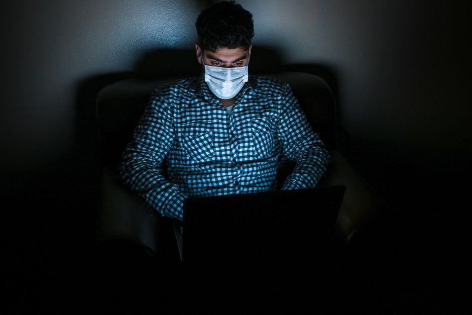 暗い部屋でラップトップを使用しているマスクを着用した男性、デジタルノマドライフスタイルを強調しています。