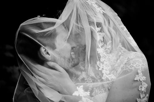 Black and white of happy bride and groom covered in veil while hugging and looking at each other on black background