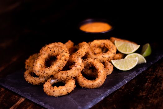 Vibrant orange salad dressing and fried brown round onion rings composing with slices of lime on wooden table in daylight