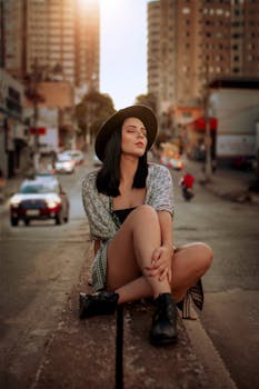 Fashionable woman in a fedora hat sits on a city street at sunset, exuding style and relaxation.