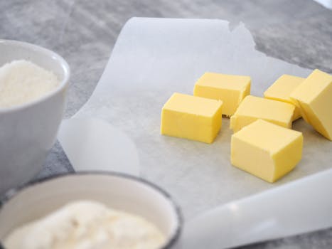 Close-up of butter cubes on parchment paper with bowls of ingredients in a kitchen setting.