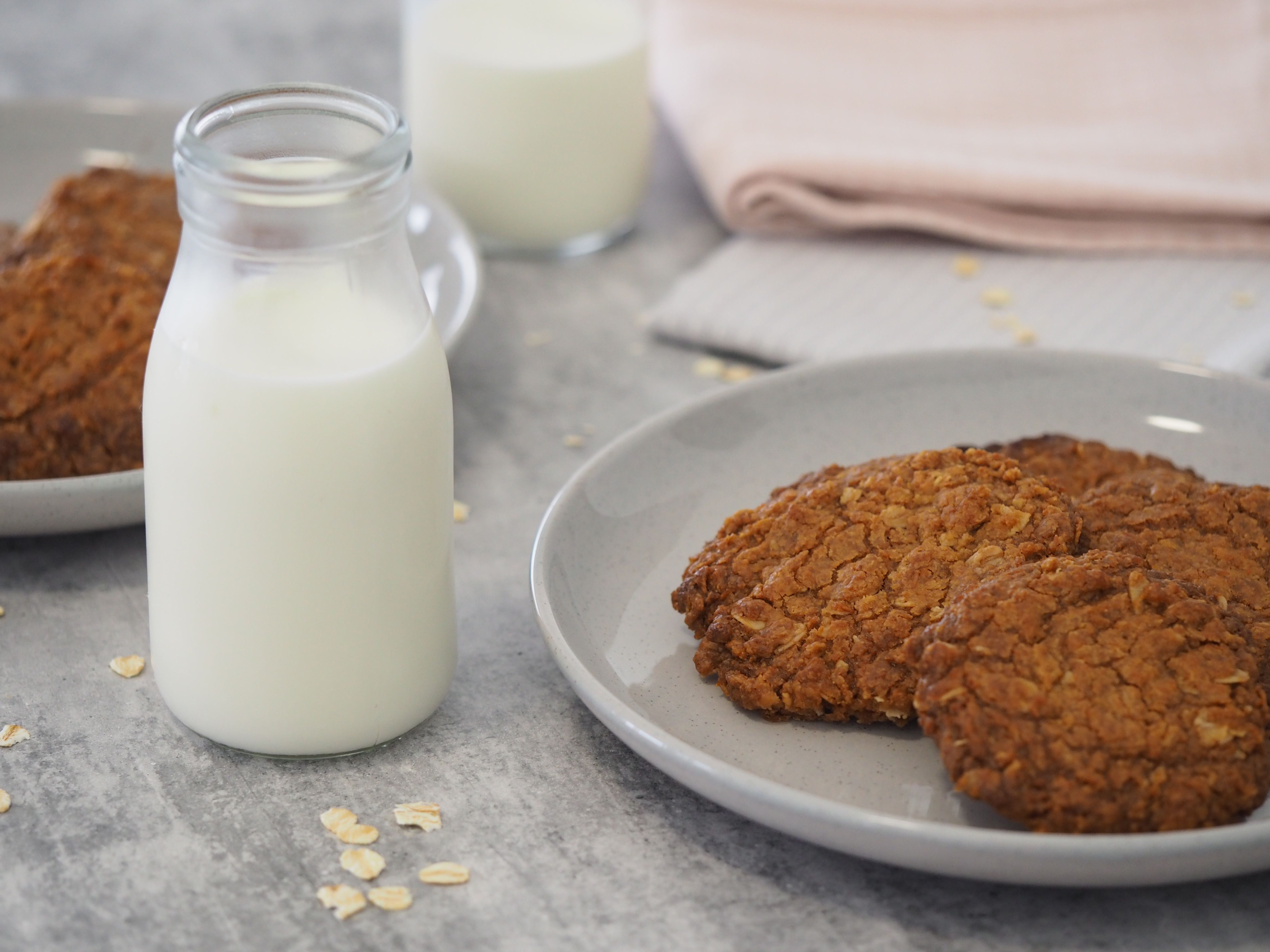 A glass jar and glass of pale, creamy oat milk on a wooden counter next to a small bowl of rolled oats and a nut milk bag.