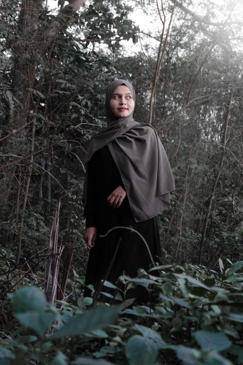 Free From below of positive young ethnic female in black dress and traditional hijab standing in forest amidst green trees and looking away Stock Photo