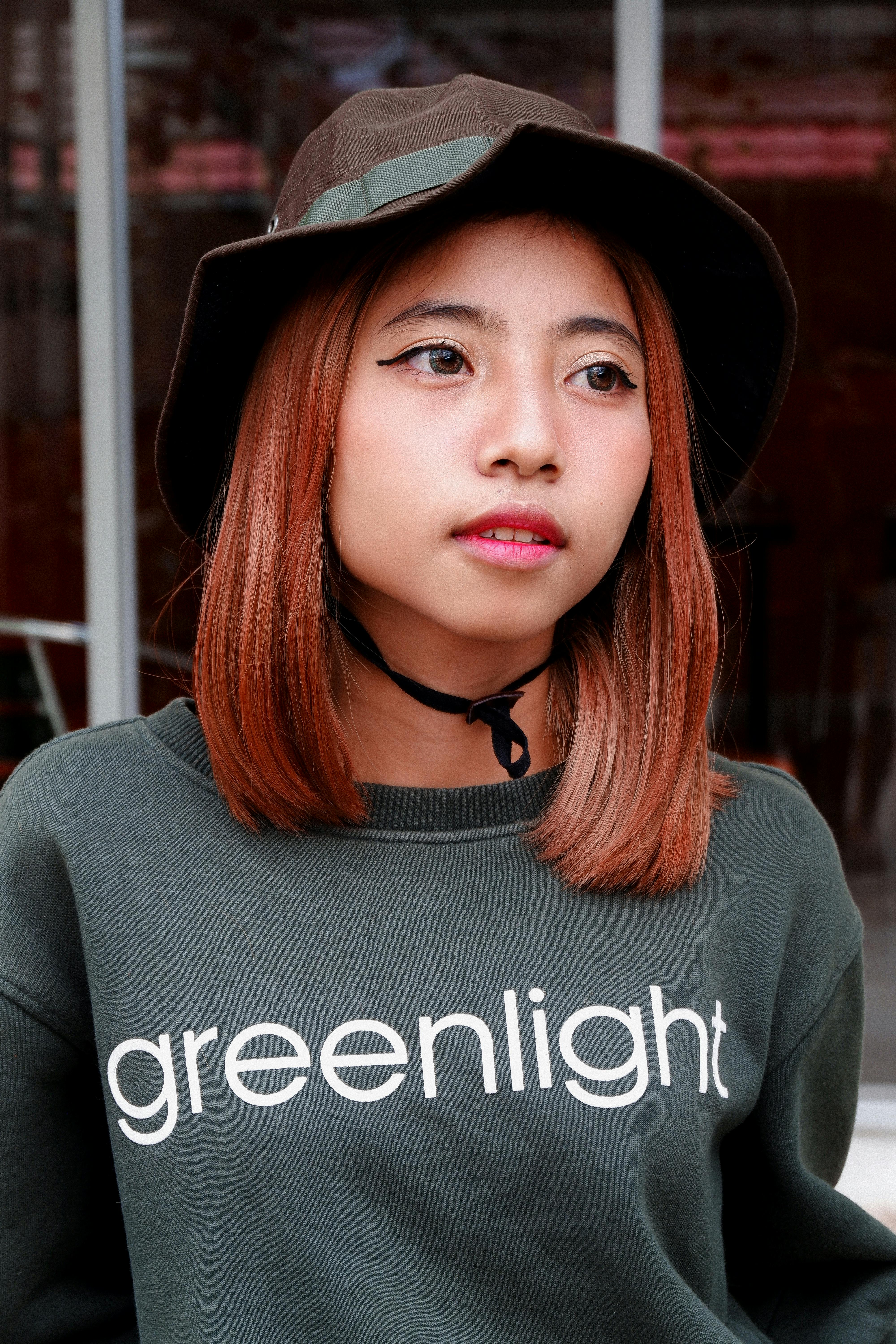 Pensive young Asian female millennial with red hair in trendy outfit standing on city street and looking away