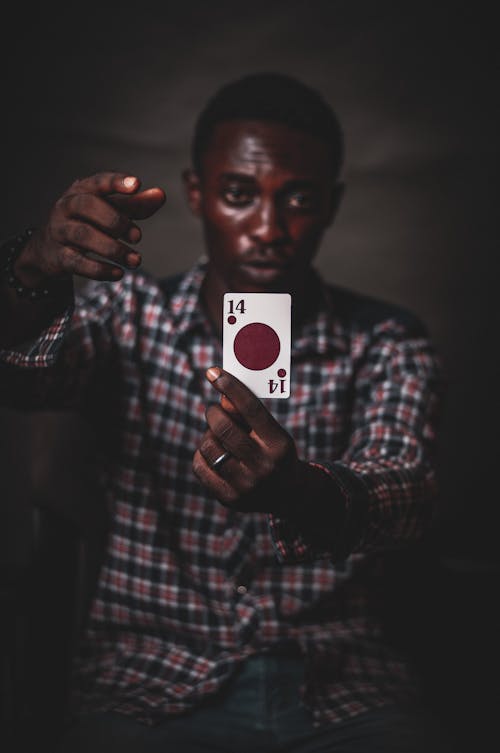 Focused young African American male in checkered shirt demonstrating playing card with red circle against dark background