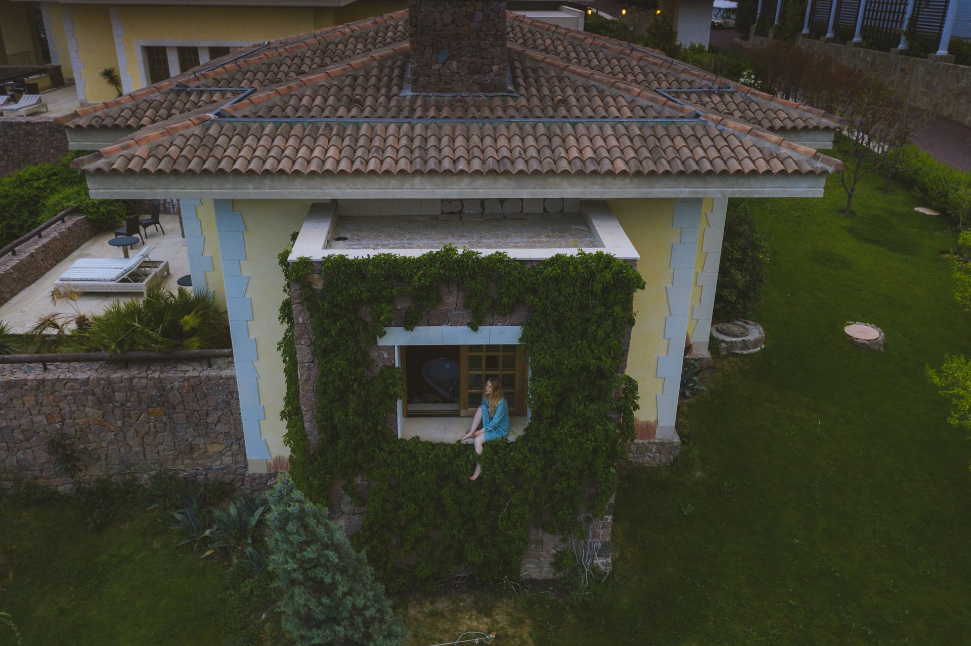 Aerial view of a woman sitting on a window sill of a vine-covered house in a garden setting.