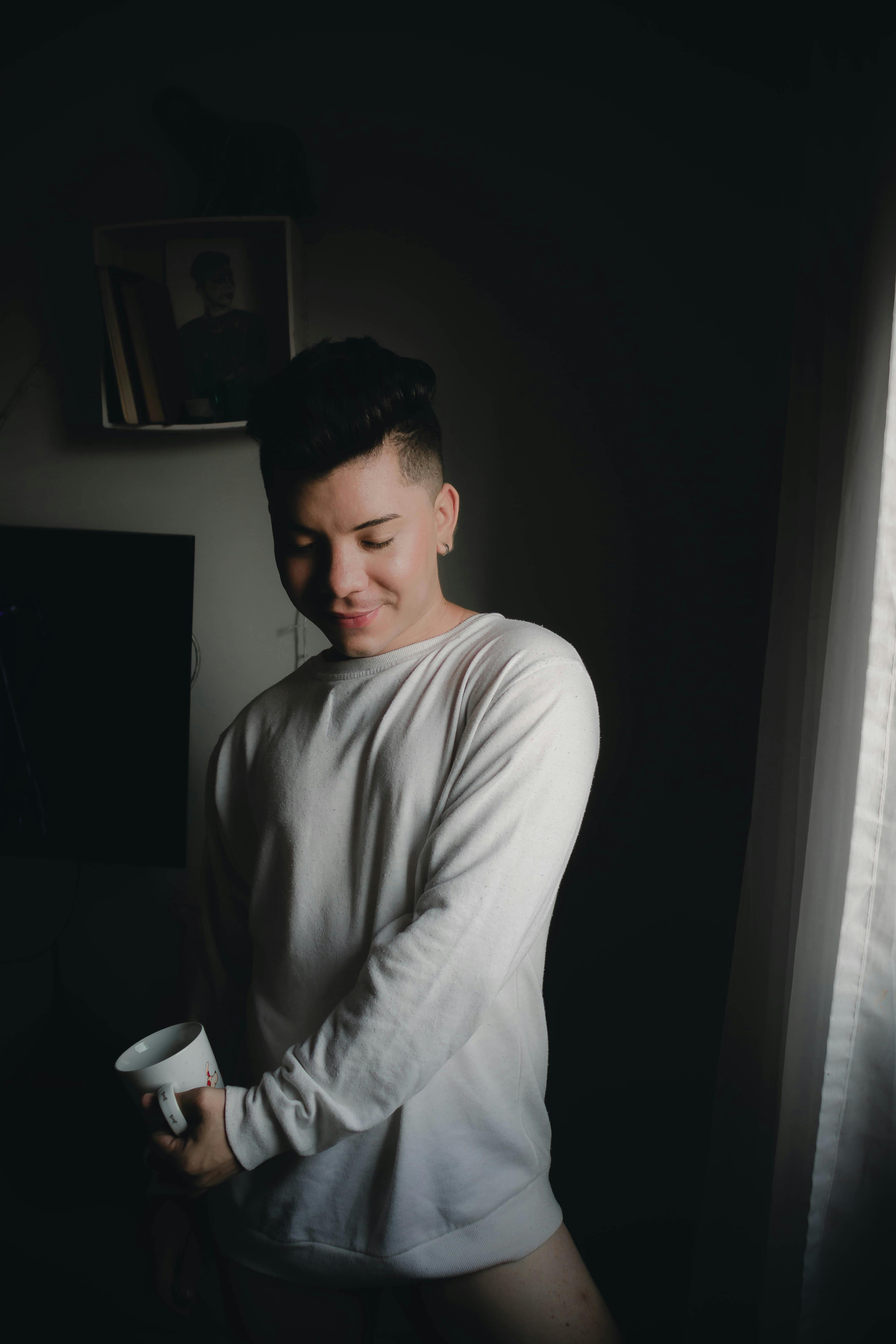 Young man in a white shirt standing by a window, holding a cup, captured in soft indoor lighting.