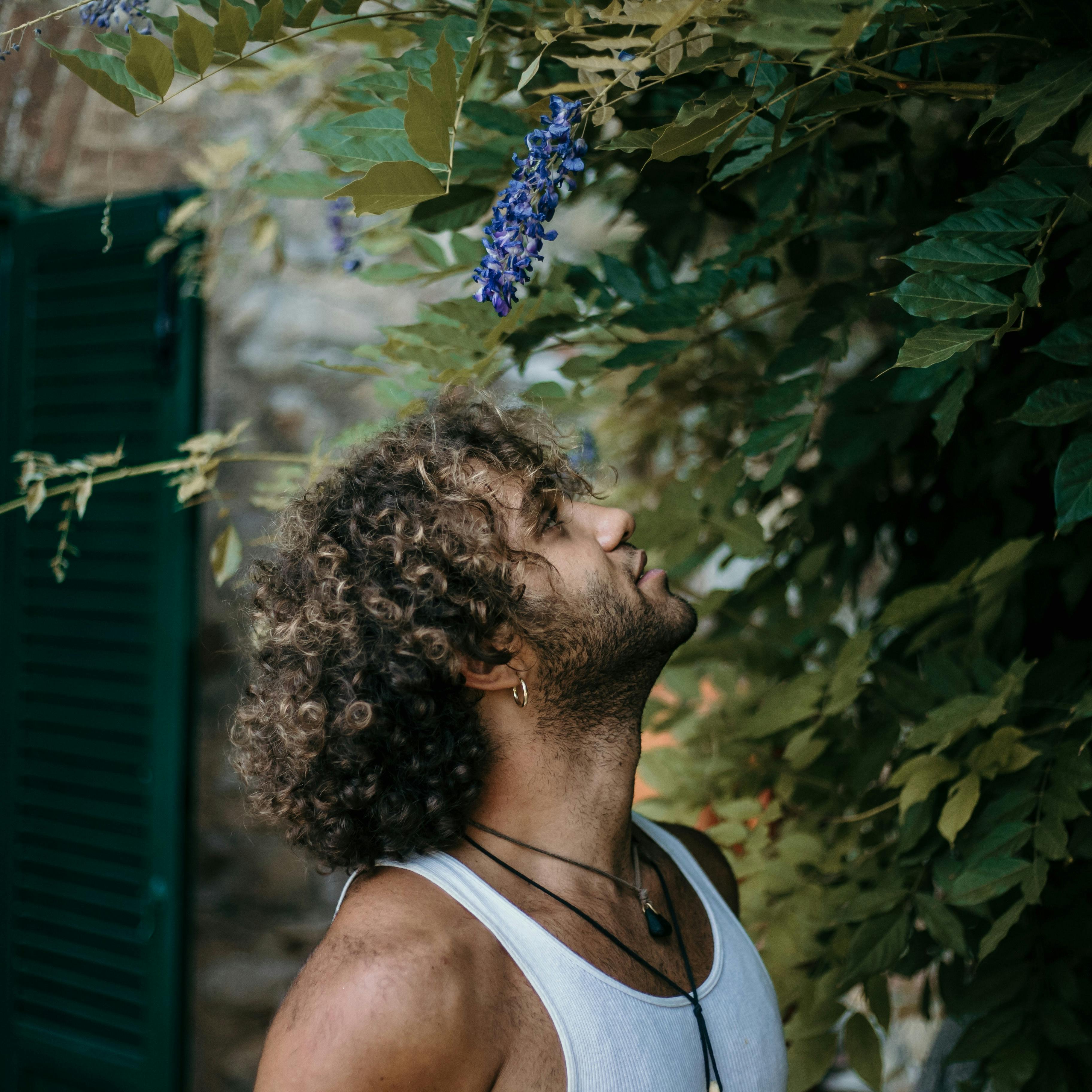 A man with curly hair looks up at purple wisteria flowers outdoors in a serene Italian setting.