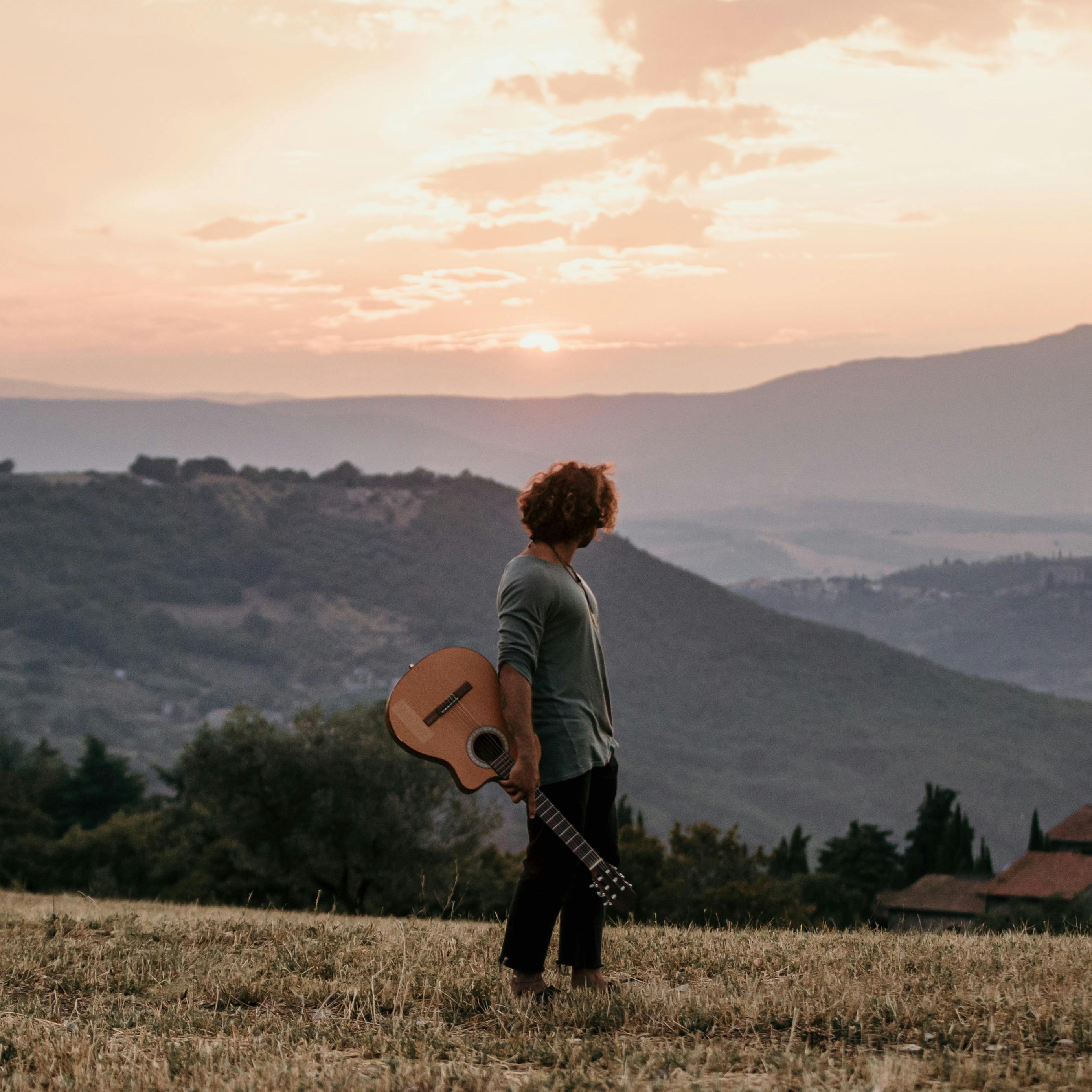 A man stands in a scenic Italian landscape holding a guitar during sunset.