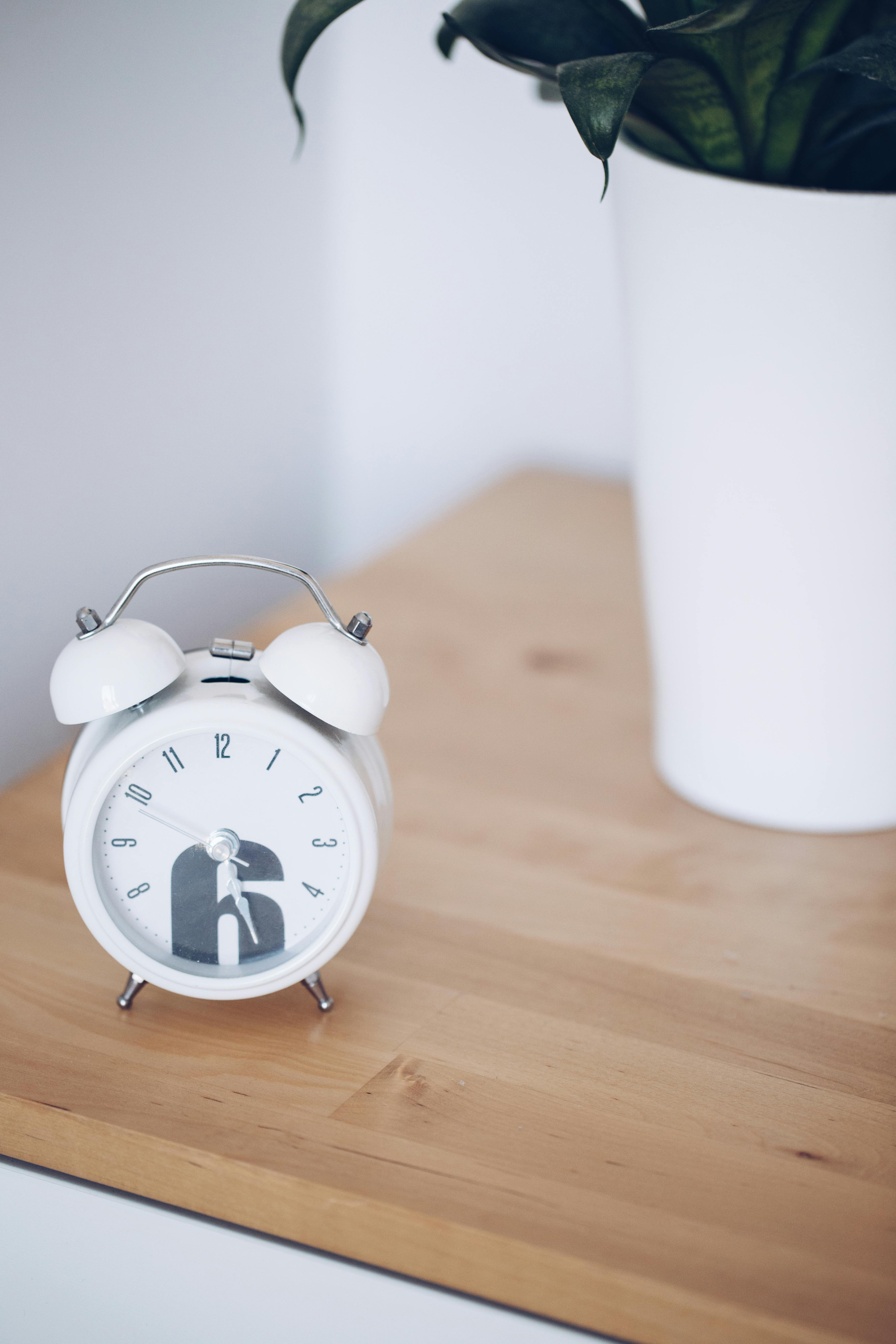 Close-up of a white alarm clock on a wooden table, next to a plant in a modern minimalist setting.