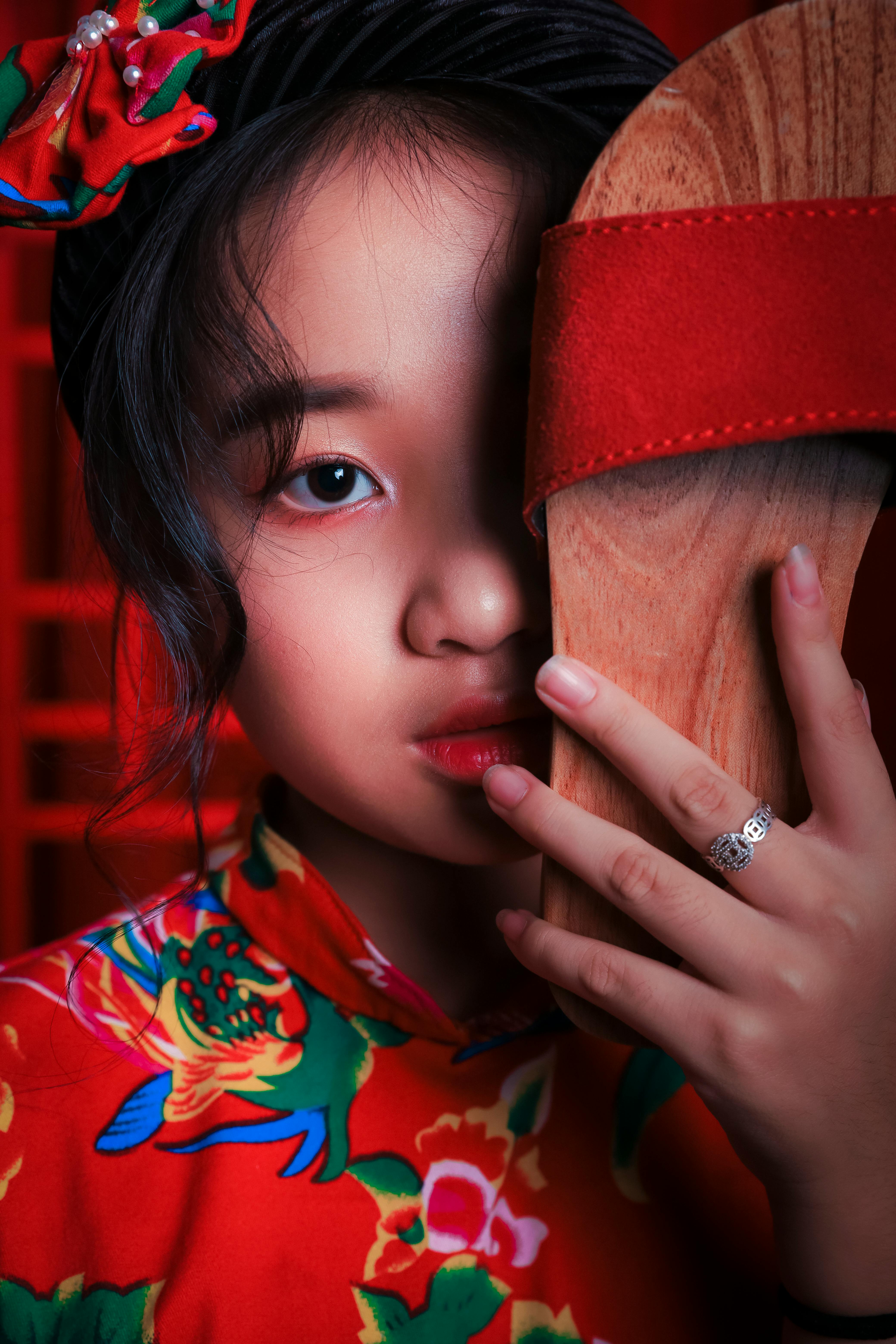 Crop trendy ethnic child in bright apparel with accessories covering half face with sandal while looking at camera