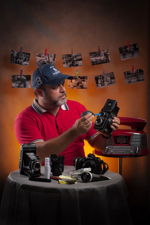 Concentrated male worker in cap applying oil to vintage film camera while sitting at table with assorted lubricant tubes and photo cameras