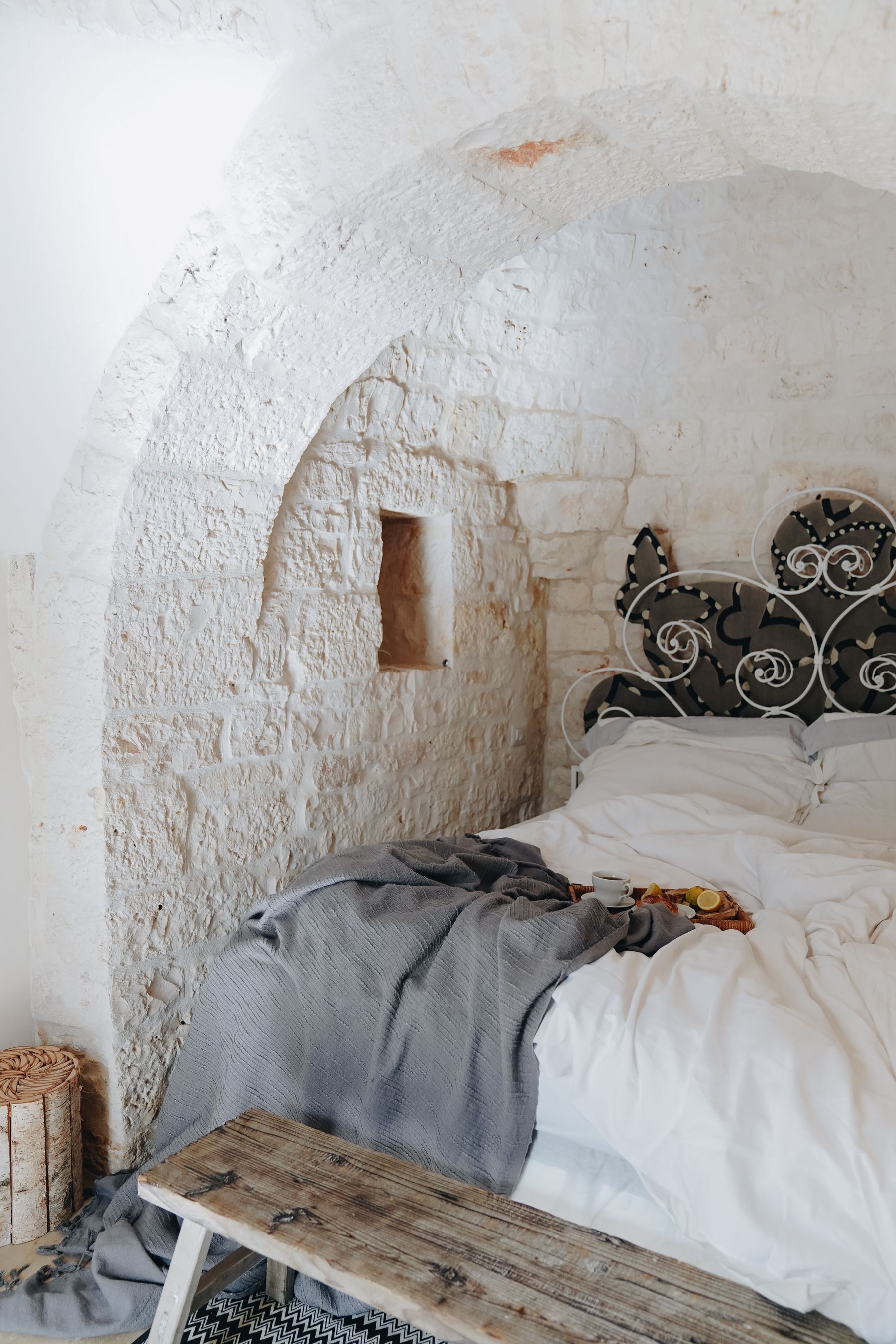 Rustic stone-walled bedroom with a black iron bed, white bedding, wood bench and warm light