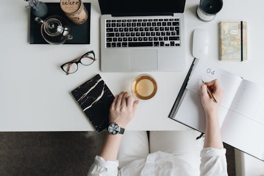Top view of a home office desk with a laptop, planner, pen, tea, and glasses, showcasing a productive workspace atmosphere.