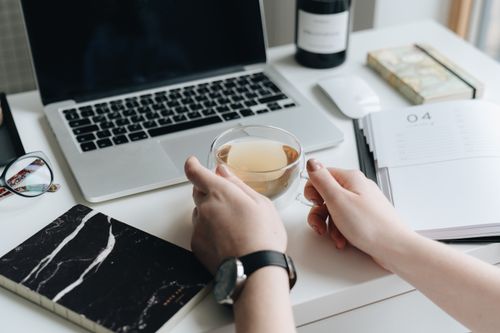 Free A person holding a tea cup at a stylish workspace with a laptop and notebook. Stock Photo