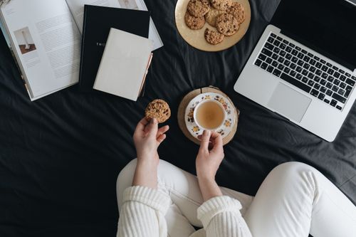 Free A cozy indoor scene of a person enjoying cookies and tea near a laptop and books on a bed. Stock Photo