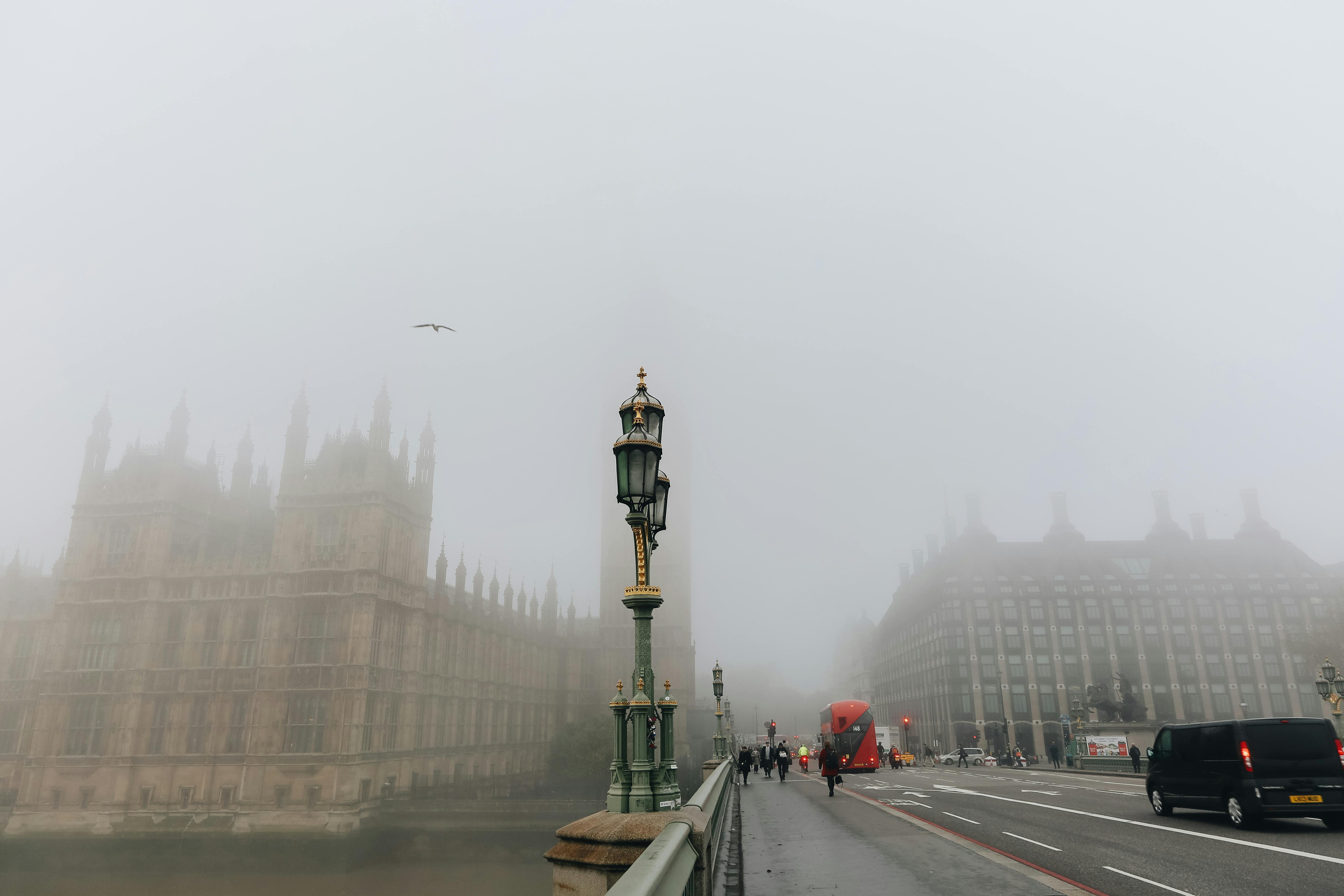 Misty morning view of Westminster Palace and Big Ben shrouded in fog, with a red double-decker bus on the road.
