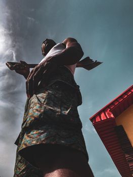 From below of anonymous African American male in shorts standing with old gardening shovel on shoulder while looking away under cloudy sky