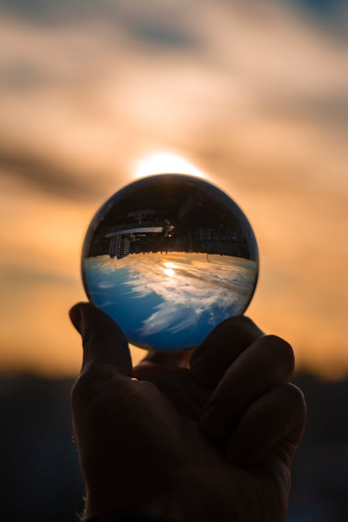 Crop anonymous person showing glass ball reflecting sky with clouds and shiny sun in twilight