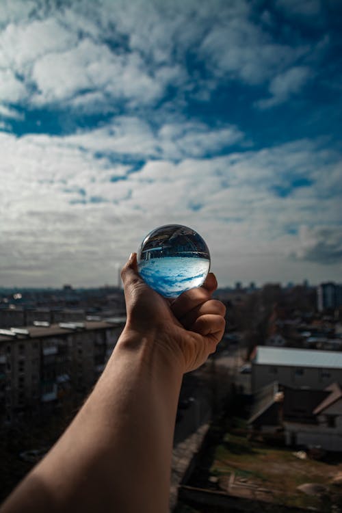 Crop unrecognizable person with reached arm showing crystal ball reflecting blue cloudy sky above city