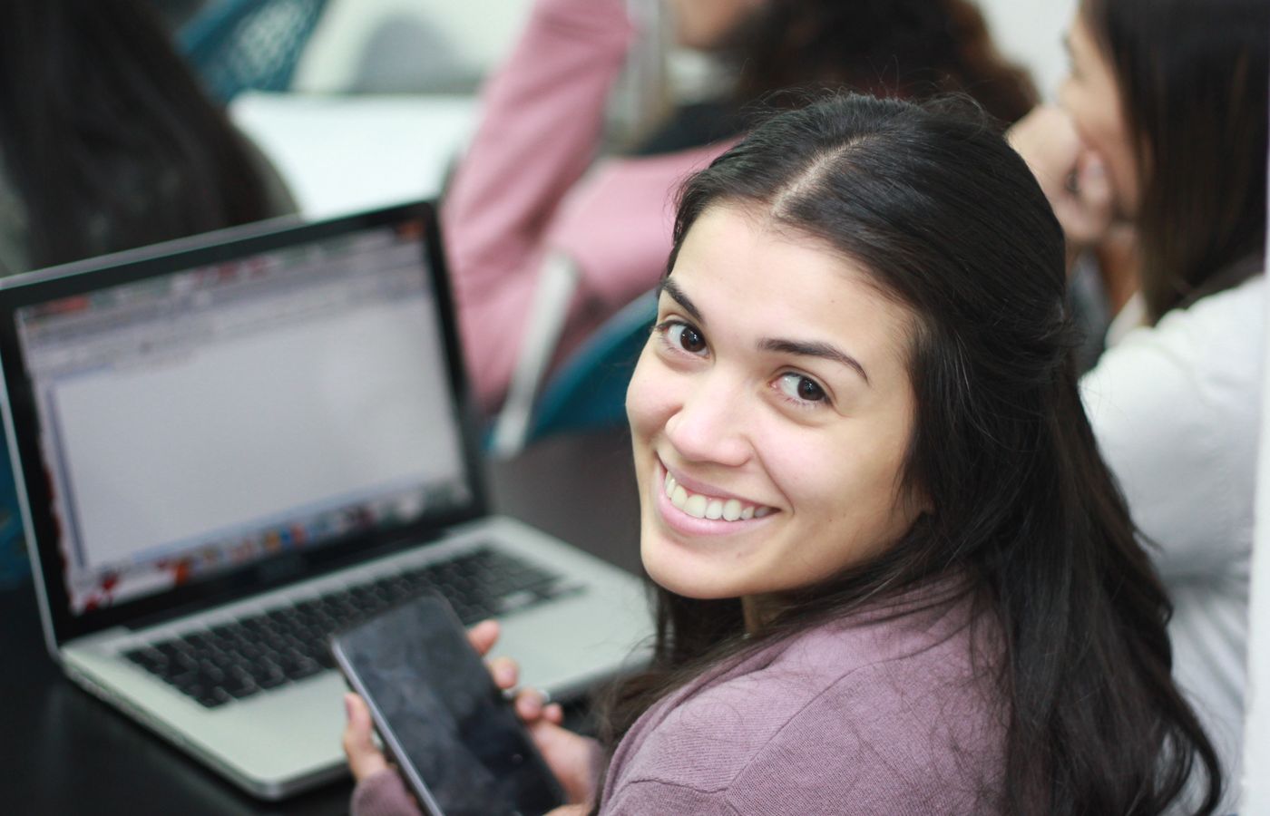 Woman smiling at camera while holding smartphone, seated next to laptop in indoor setting.