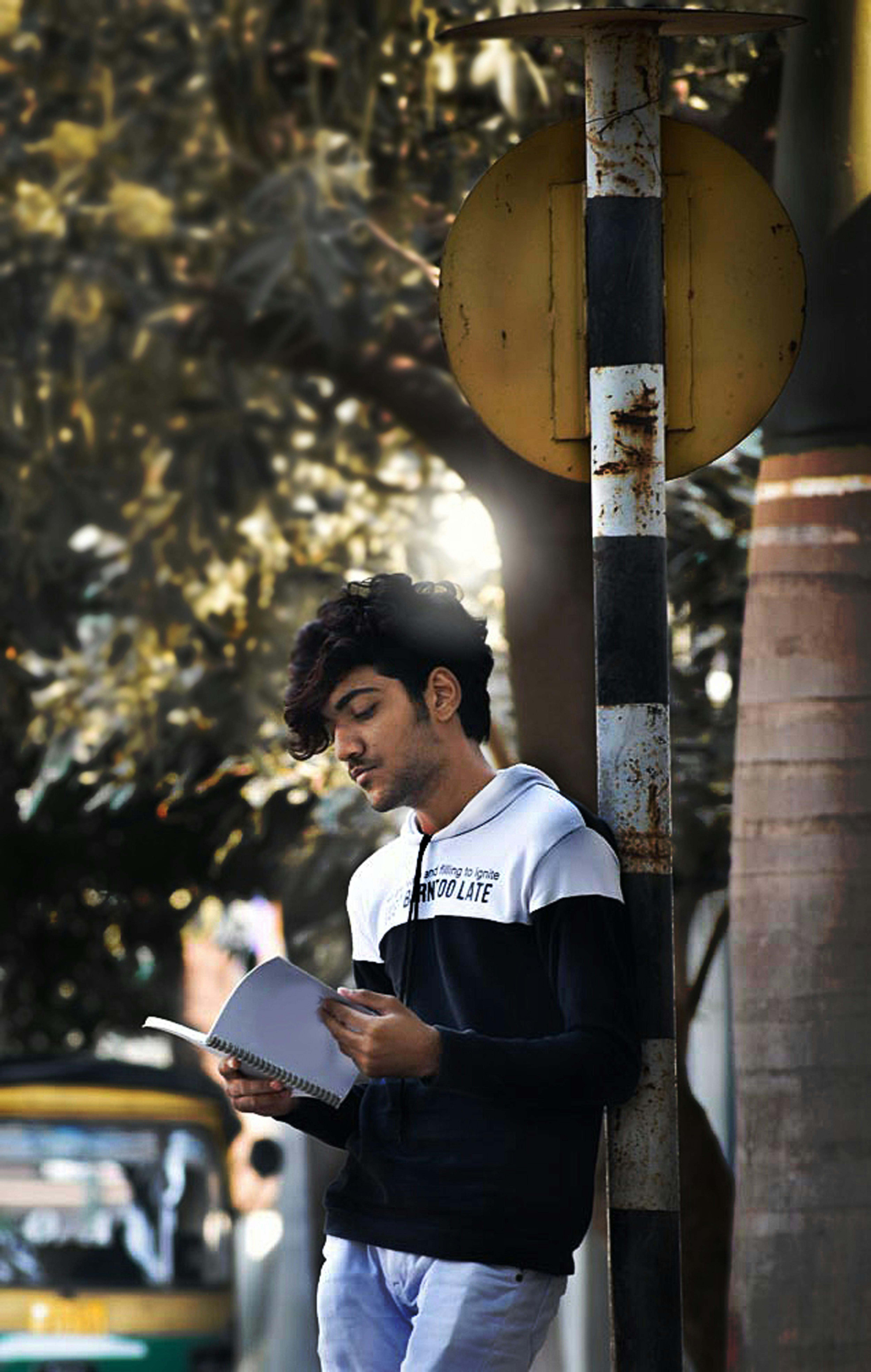A young man leans on a pole, reading a notebook outdoors in casual attire, India.