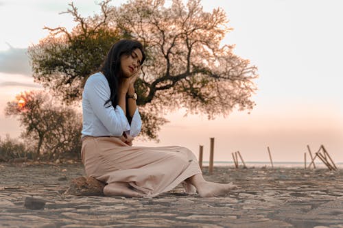 Side view of young ethnic woman with sleep gesture sitting on tree trunk on sandy shore near ocean at sunset