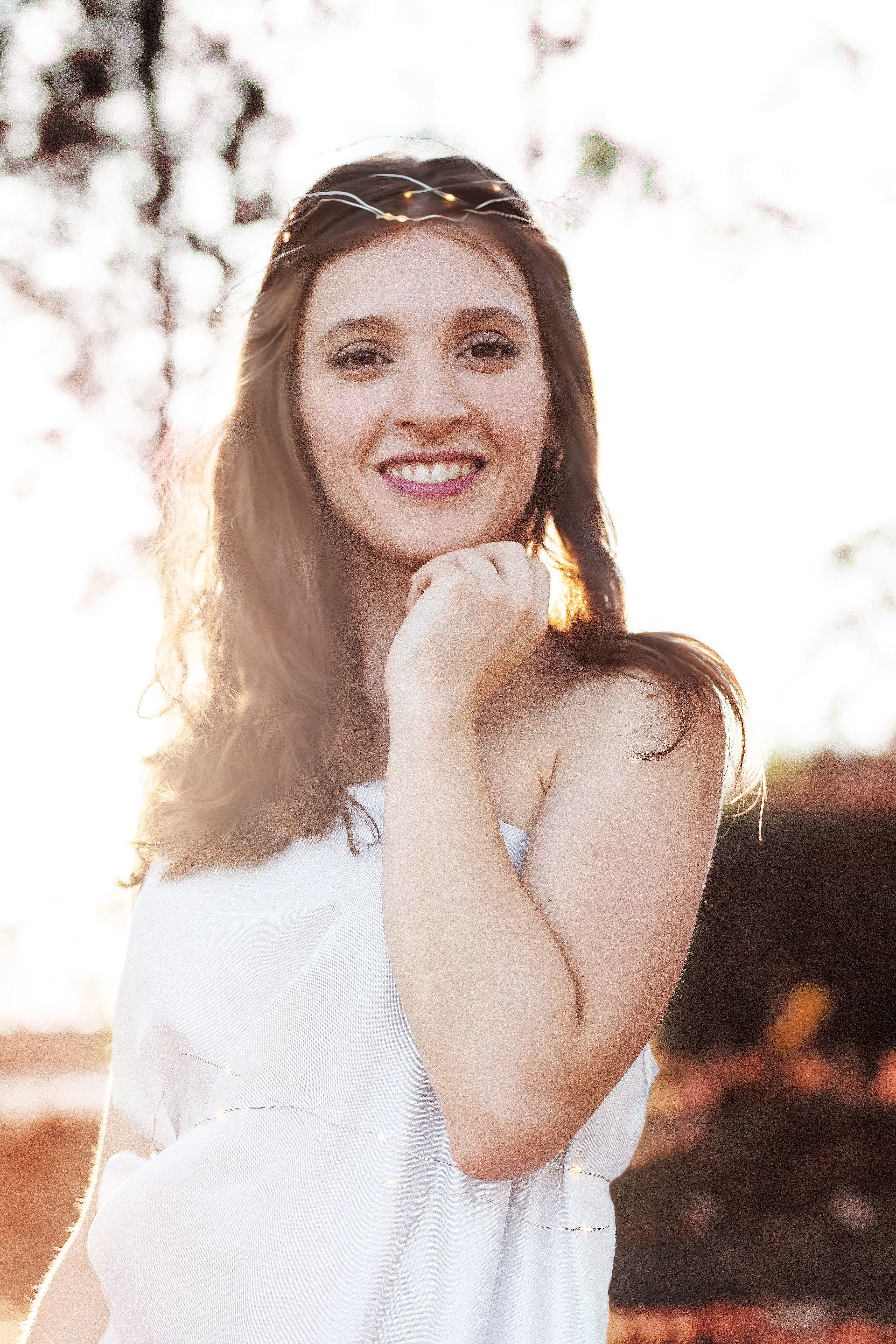 From below of happy trendy female in white clothes touching chin while standing under sky and looking at camera in back lit