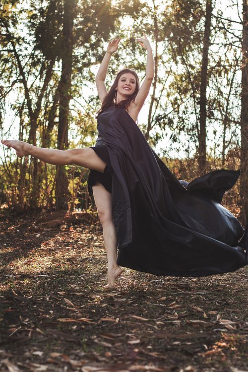 Free Young cheerful ballet dancer in black wear standing with reached leg and raised arms while dancing near trees in summer Stock Photo