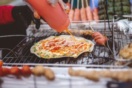 Close-up of a hand applying sauce to a pizza on a grill, highlighting street food preparation.