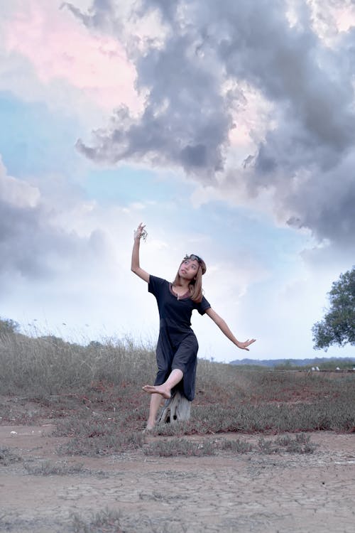 Serious young female in casual dress sitting on stump in field under cloudy sky