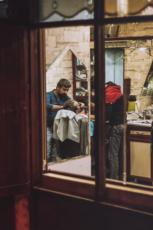 Through window glass of concentrated bearded male barber trimming beard of client in barbershop