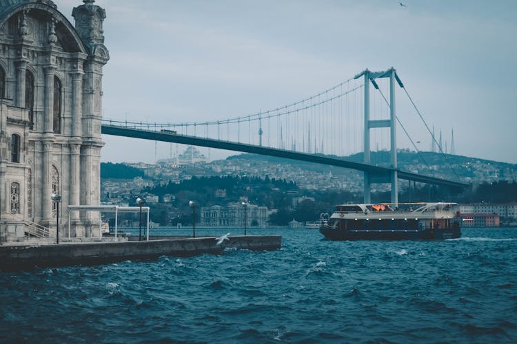 Cruise Boat Floating On River Near Suspension Bridge And Old Mosque